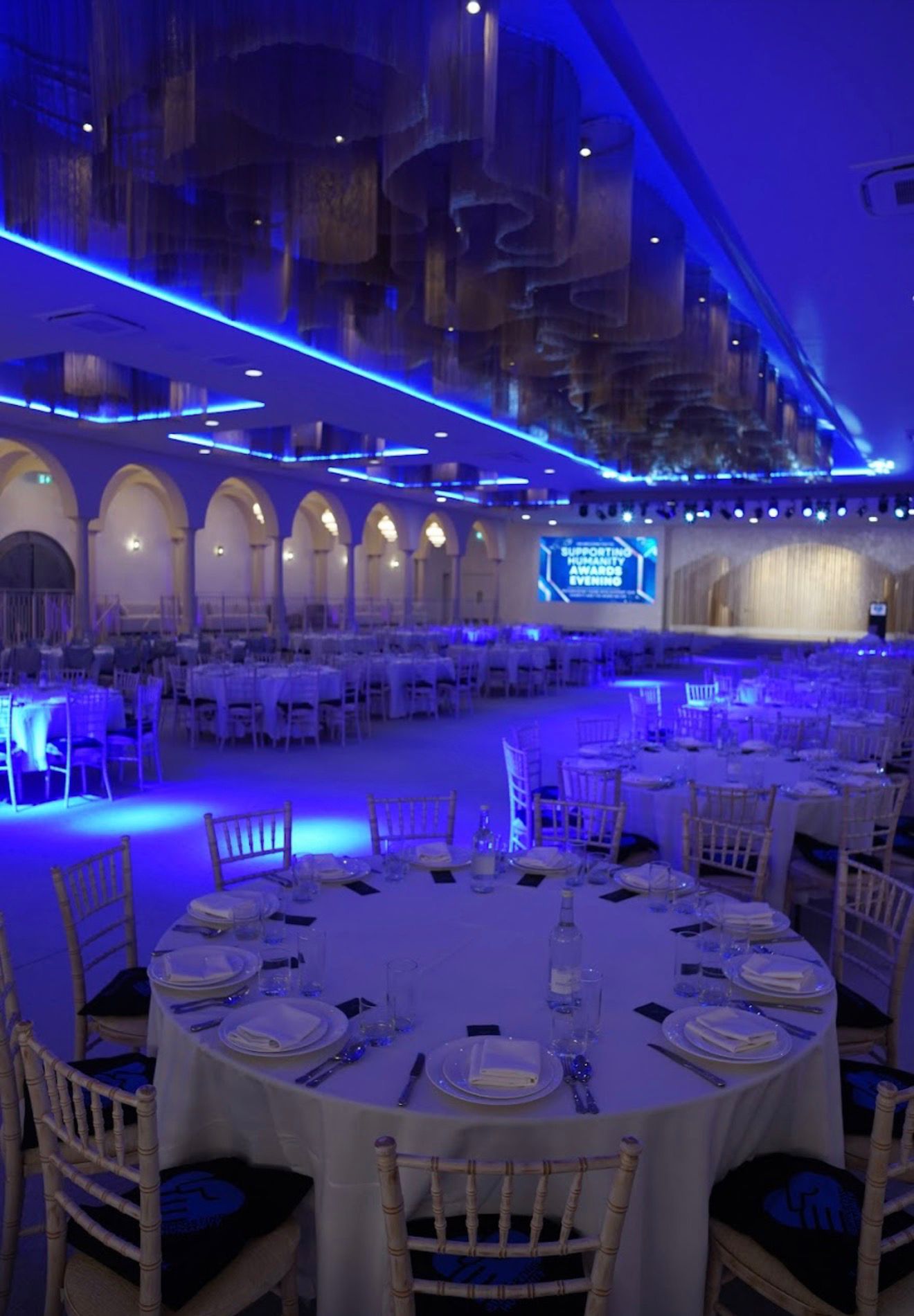 Blue-lit gala hall with large screen and round tables